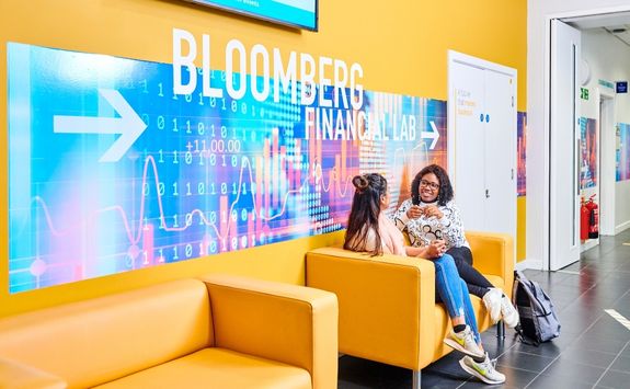 Two students sitting on a yellow sofa in front of a large colourful sign reading "Bloomberg Financial Lab" with an arrow pointing right